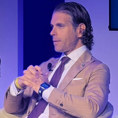 Domenico De Rosa in giacca cammello, camicia e cravatta seduto su una poltrona, fotografato di profilo e con le mani incrociate.