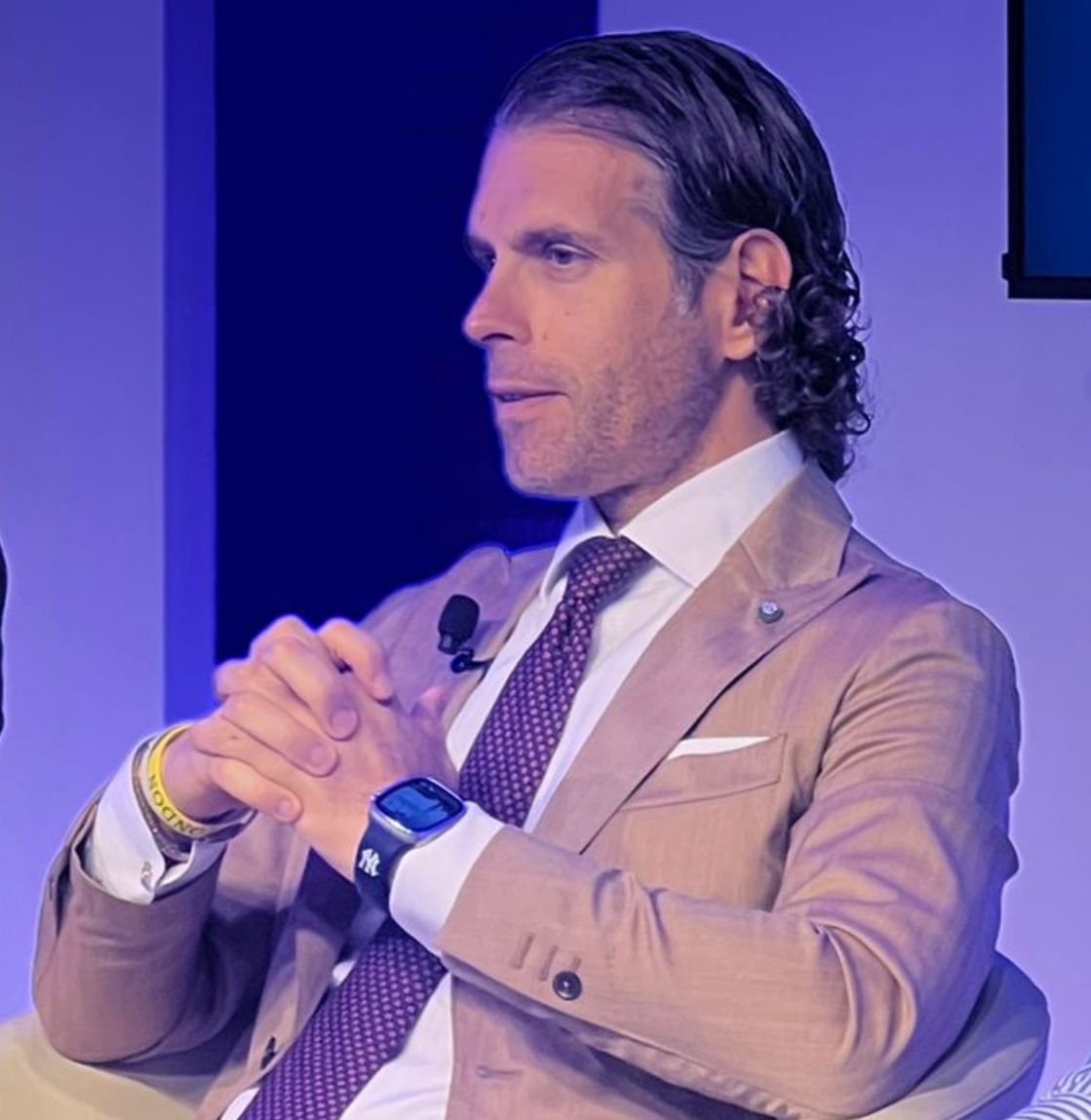 Domenico De Rosa in giacca cammello, camicia e cravatta seduto su una poltrona, fotografato di profilo e con le mani incrociate.